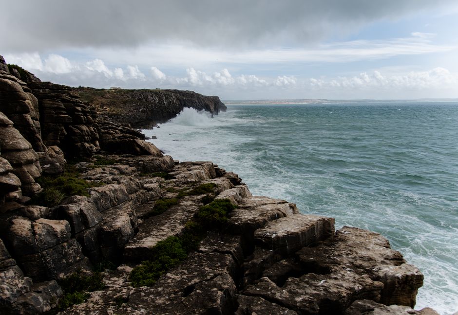 Dramatic view of a rocky coastline with waves crashing against the cliffs under a cloudy sky.