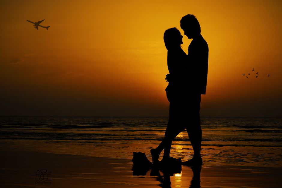 Silhouette of a couple embracing at the shore during sunset with an airplane overhead.