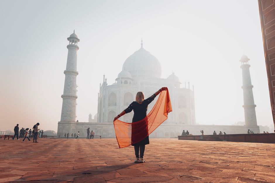A woman stands before the majestic Taj Mahal at sunrise, gracefully holding a red scarf.