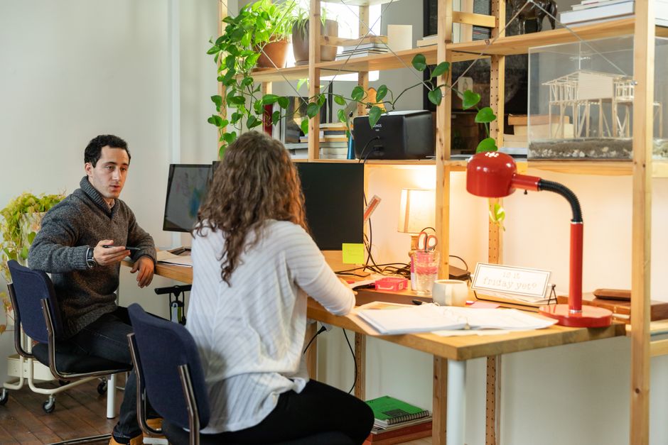 Two colleagues discuss work at a modern office desk with multiple monitors and greenery.