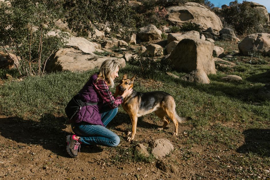 A woman kneels to pet her dog in a rocky, sunny outdoor landscape.