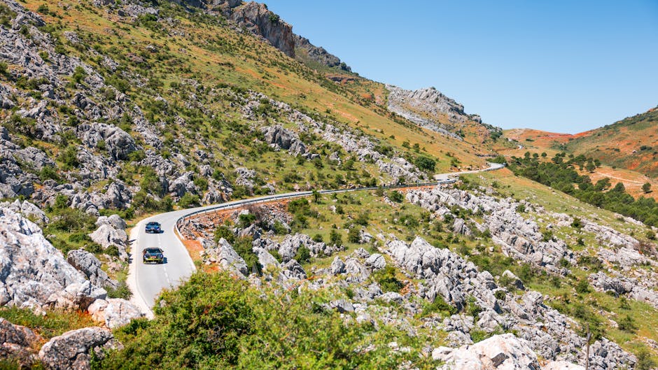 Two cars navigate a winding road through a scenic mountain landscape under clear blue skies.