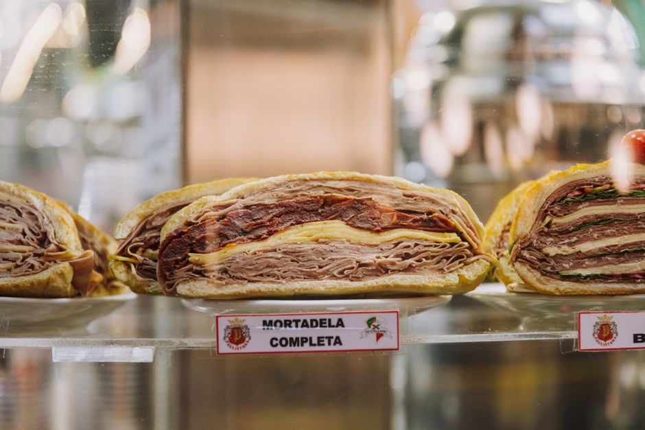 Close-up of assorted sandwiches with mortadela, cheese, and greens on display at a deli or bakery.