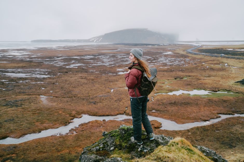 A woman stands on a rocky outcrop, gazing over a foggy, cold landscape with a backpack and warm clothing.