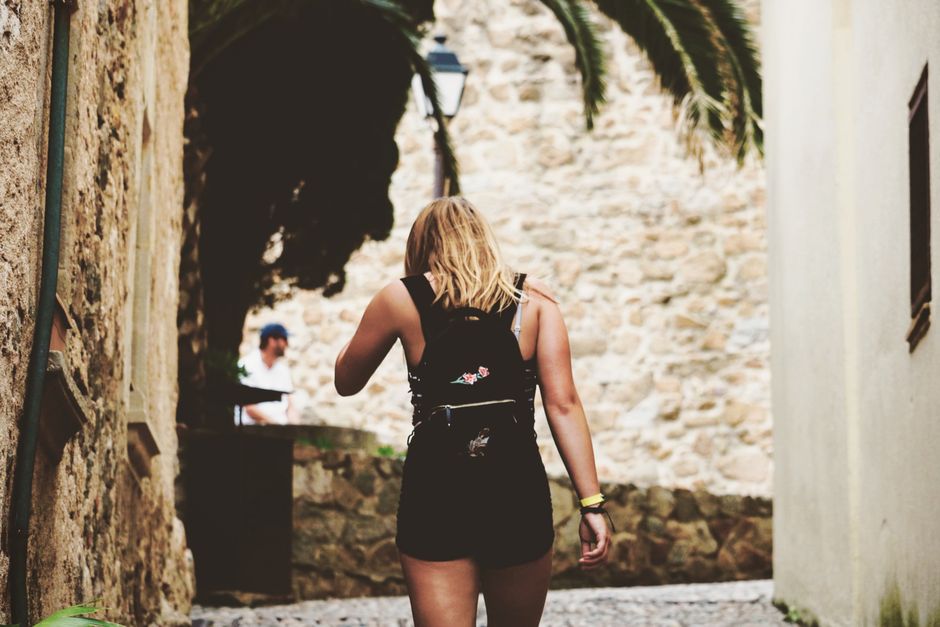 Blonde woman strolls through a picturesque stone alleyway on a sunny day, wearing a fashionable black outfit.