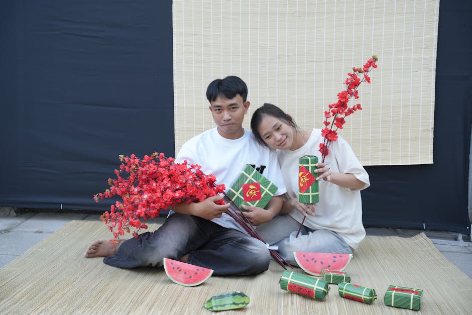 Asian couple with Tet decorations, holding red blossoms and gifts, smiling indoors.